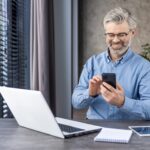 Freelancer d'âge moyen satisfait à l'aide de téléphone mobile par ordinateur avec ordinateur à côté de la fenêtre avec